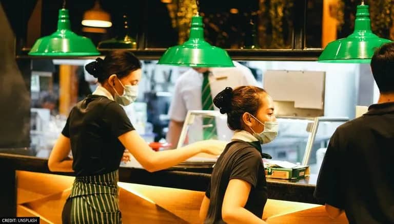 Two female wait staff, masked, wearing striped aprons, line up at the pass. There's another male server to their right. The pass overhead lights are a lovely green, old-style metal shade.