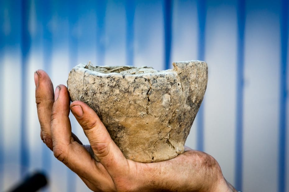 Against a blue & white striped background, an ancient ceramic pot damaged, broken but still mainly intact is cupped in a man's hand. His fingers and nails show ingrained dirt, perhaps from the earth that surrounded this item?
