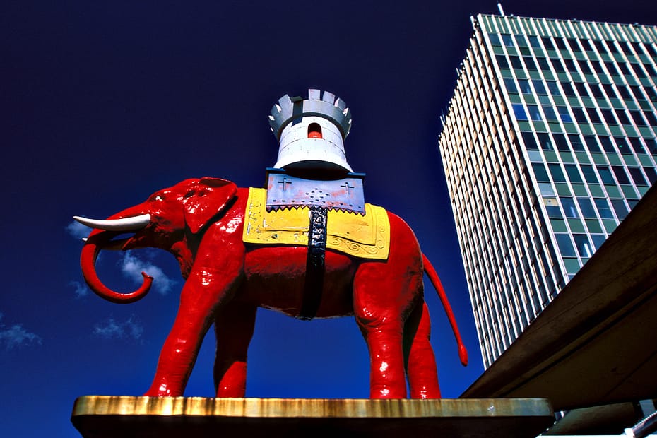 Statue of the Elephant & Castle (salvaged from the Victorian pub) set against an anonymous tower block. Red body, yellow rug over the back and then a castellated white and blue tower on top.