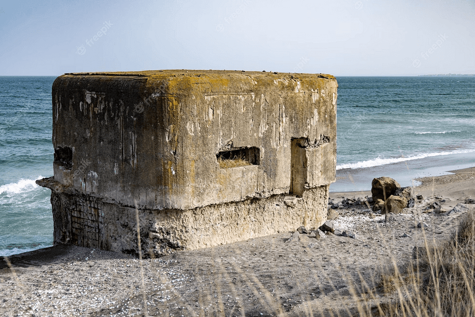 An old WWII concrete bunker; moss and growth on the roof, slowly rotting away, with slit windows against a blue sea, waves coming in slowly, the foreground is beach and wild grasses.