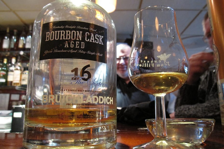 A nearly empty bottle of Bruichladdich 16 Year Old Bourbon Cask whisky rests on a pub bar, next to a tapered glass with some of the Scotch still in it. Behind in the background is a man in glasses.