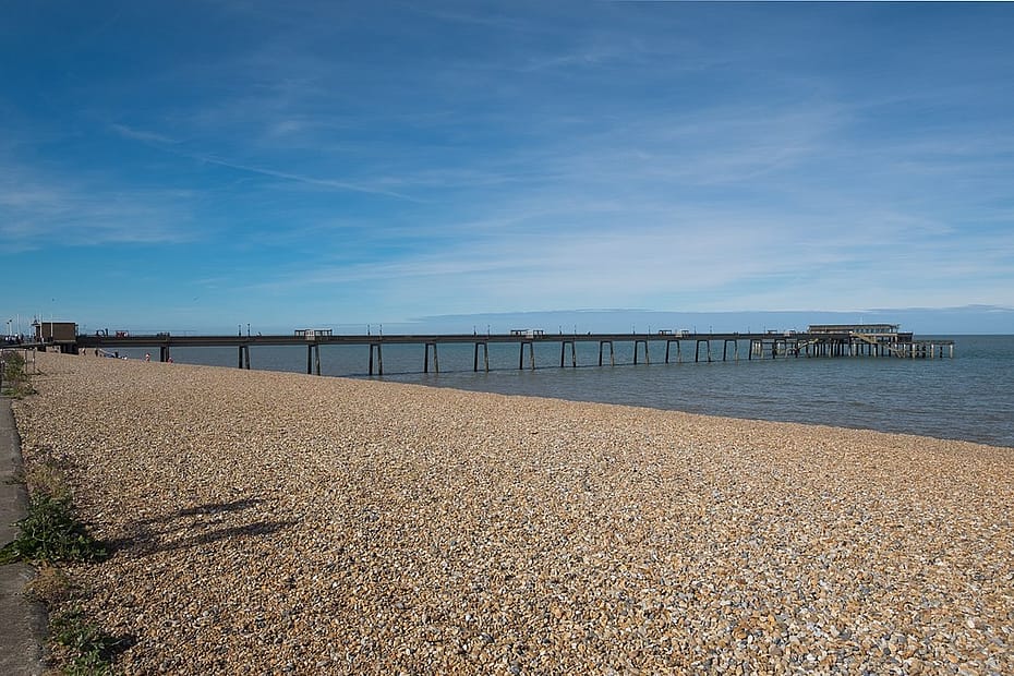 A pier running left t right into the blue-grey sea. single beach in the foreground. A small sea wall at the very left edge of the picture.