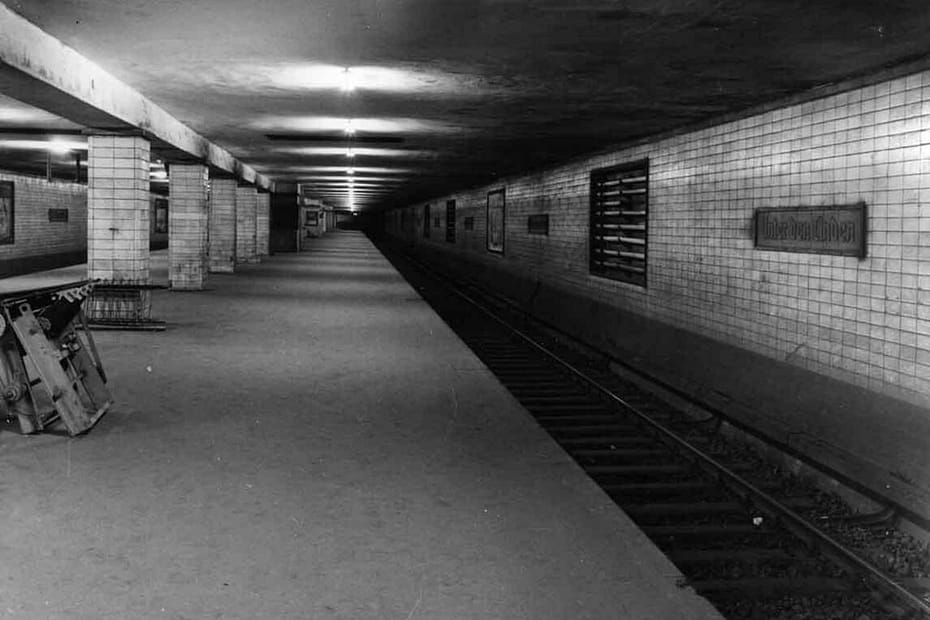 A "ghost" tube station underneath Berlin Unter Den Linden showing two lines converging into one in the foreground. Some lights but lots of shadows and gloom remain. Old crates litter the platform, and a station name sign in old German script is on the tiled wall.