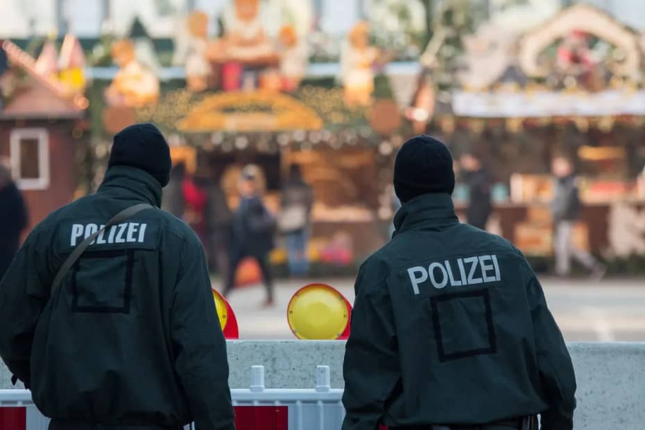 Two black-clad armed German police officers with balaclavas facing away from the camera are standing behind a waist high wall or barrier looking out over a street, the background makes it look like this is Christmas time.