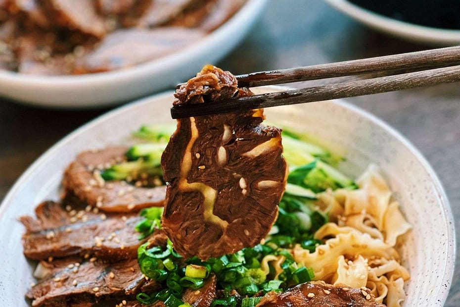 Slices of beef in a circles, on a wjite plate with some noodles to the side and chopsticks holding greens above it.